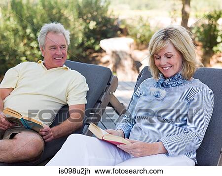 Stock Image - Mature couple reading on loungers in garden. Fotosearch