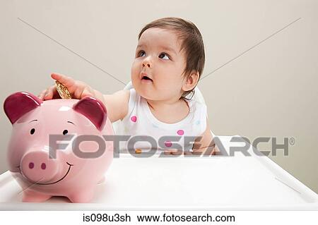 Stock Image - Baby boy sitting in highchair with piggy bank and coins. Fotosearch