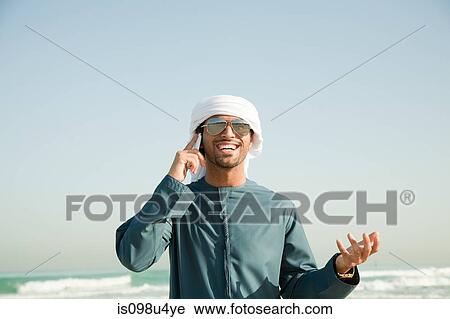 Stock Image - Middle Eastern man using mobile phone on the beach. Fotosearch
