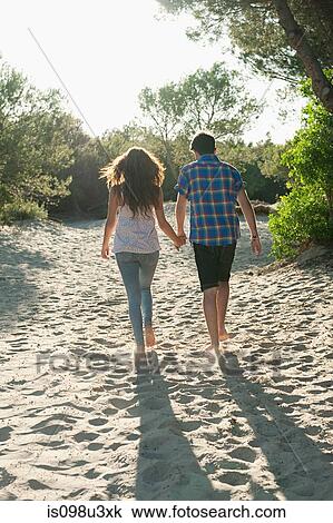 Stock Image - Young couple walking across sand, rear view. Fotosearch