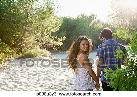 Stock Image - Young couple walking across sand, rear view. Fotosearch