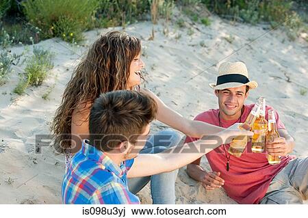 Stock Image - Young friends sitting on sand with beer. Fotosearch