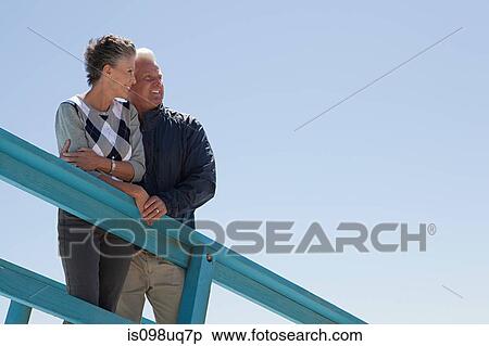 Stock Photography - Mature couple by railing against blue sky. Fotosearch