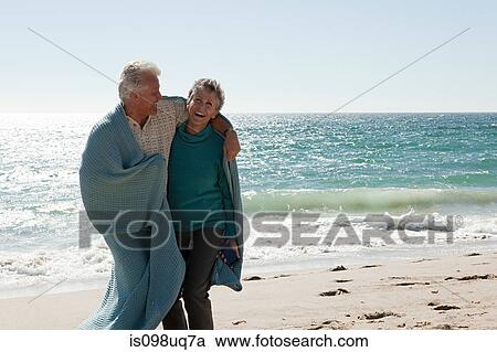 Stock Image - Mature couple on beach wrapped in blanket. Fotosearch
