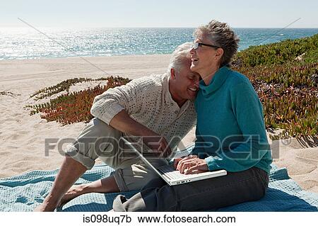 Stock Image - Mature couple using laptop at the beach. Fotosearch