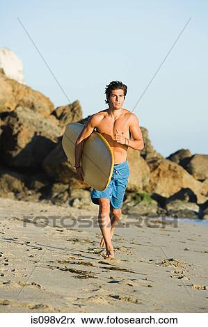 Stock Photo - Young man running on beach with surfboard. Fotosearch