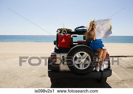 Stock Image - Young woman sitting on vehicle looking at map. Fotosearch