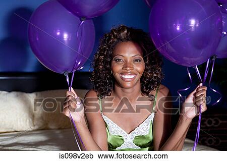 Stock Image - Young woman on bed, holding balloons. Fotosearch