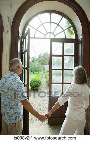 Stock Image - Mature couple holding hands by french doors. Fotosearch