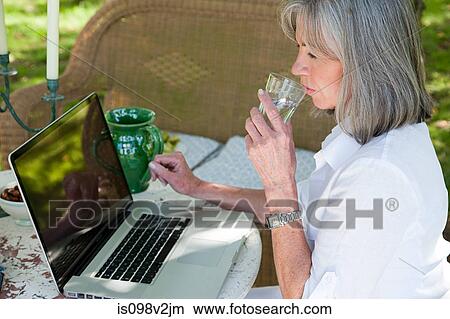Picture - Mature woman using laptop drinking water. Fotosearch