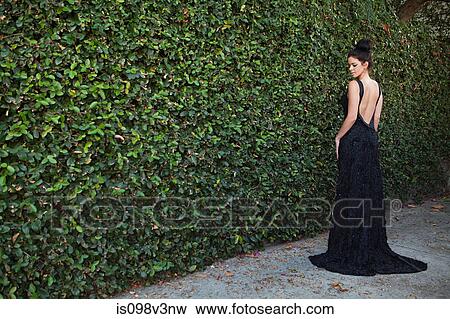 Stock Photo - Young woman wearing black evening dress, standing by hedge. Fotosearch