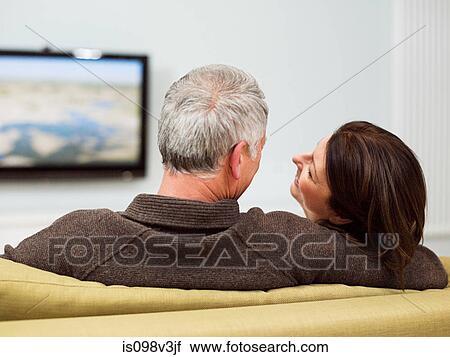 Stock Image - Mature couple sitting on sofa watching television. Fotosearch
