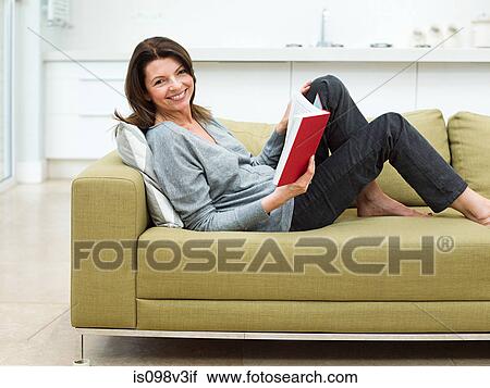 Stock Image - Mature woman sitting on sofa reading book. Fotosearch