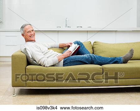 Picture - Senior man sitting on sofa reading book. Fotosearch