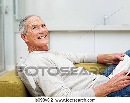Stock Image - Senior man sitting on sofa reading book. Fotosearch