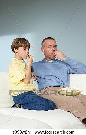 Stock Image - Father and son watching television eating popcorn. Fotosearch