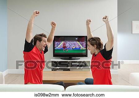 Picture - Two young men wearing football shirts watching football on television. Fotosearch