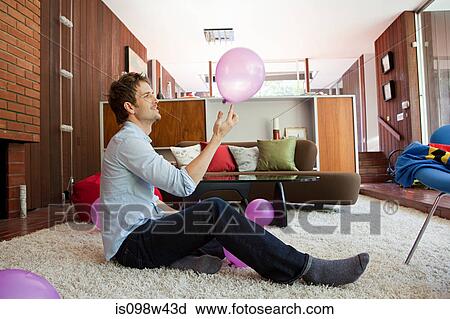 Stock Image - Man sitting on living room floor playing with pink balloon. Fotosearch