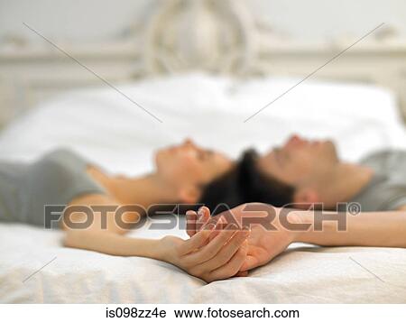 Stock Image - Young couple lying on bed holding hands. Fotosearch