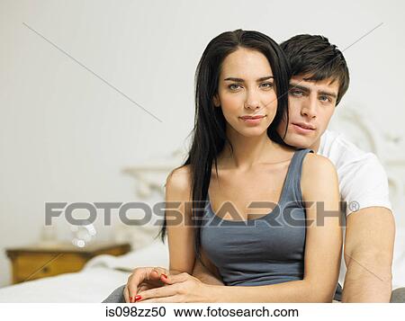 Stock Image - Young couple sitting on bed, portrait. Fotosearch