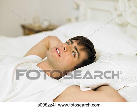 Stock Image - Young man lying on bed with hands behind head. Fotosearch