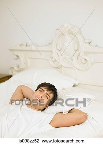 Stock Image - Young man lying on bed with hands behind head. Fotosearch