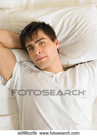 Stock Image - Young man lying on bed with hands behind head, portrait. Fotosearch