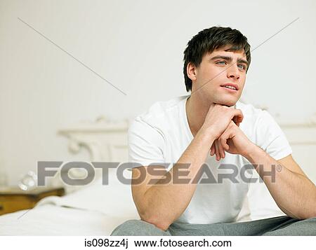 Stock Image - Young man sitting on bed, portrait. Fotosearch