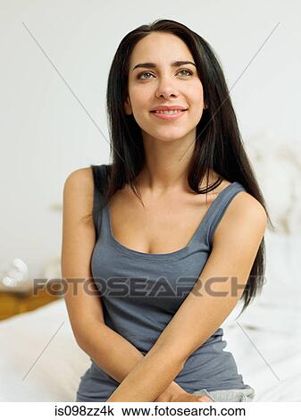 Stock Image - Young woman wearing grey vest sitting on bed, portrait. Fotosearch