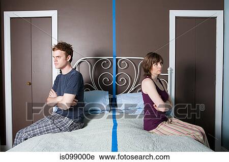 Stock Image - Young couple sitting on bed separated by blue line. Fotosearch