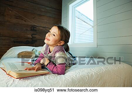 Stock Image - Young girl lying on bed reading book. Fotosearch