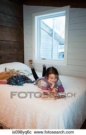 Stock Image - Young girl lying on bed reading book. Fotosearch