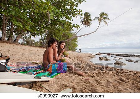 Stock Photo - Young couple sitting on beach drinking coconut milk. Fotosearch