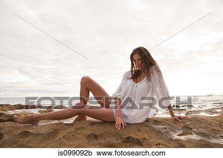 Stock Image - Young woman wearing white top sitting on sand, portrait. Fotosearch