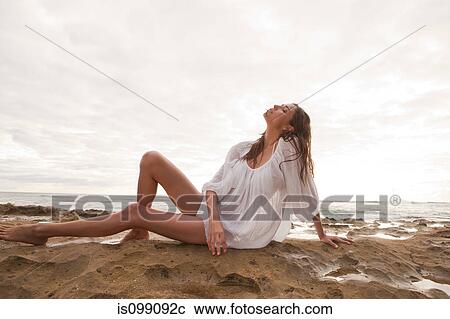 Stock Image - Young woman wearing white top sitting on sand, portrait. Fotosearch