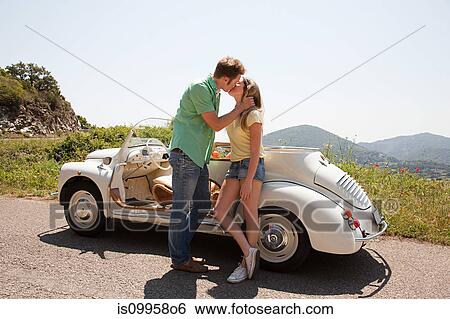 Stock Photograph - Young couple by convertible car kissing. Fotosearch