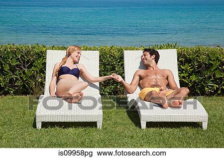 Stock Image - Young couple on sun loungers holding hands. Fotosearch