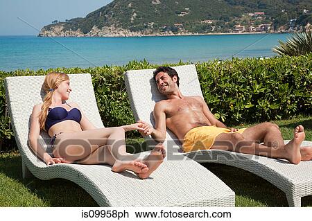 Stock Image - Young couple on sun loungers, holding hands. Fotosearch