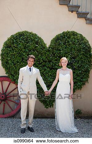 Picture - Newlyweds by heart shaped bush holding hands. Fotosearch
