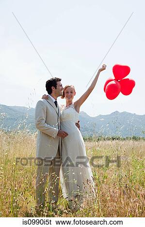 Stock Photograph - Newlyweds in field with red heart shape balloons. Fotosearch