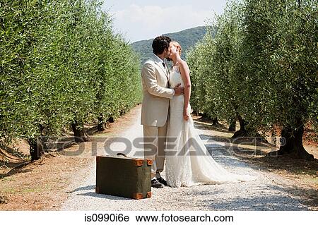Stock Image - Newlyweds kissing on country road with suitcase. Fotosearch