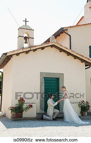 Stock Photo - Newlyweds outside church, groom on one knee. Fotosearch