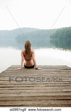 Stock Photo - Young woman sitting on pier looking at lake. Fotosearch