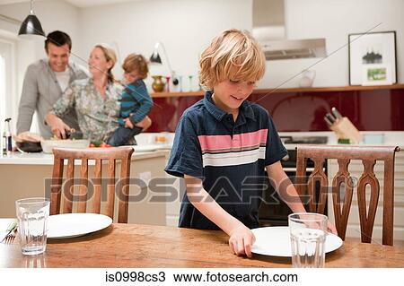 Stock Image - Son helping to lay table with parents and brother visible behind in kitchen. Fotosearch