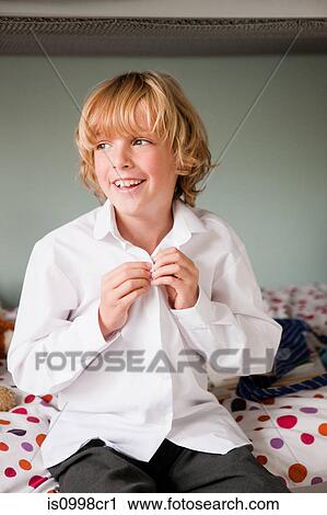 Stock Image - Young boy doing up the buttons of his school shirt while sitting on his bed. Fotosearch