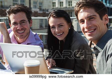 Stock Image - Three people smiling at the camera while using a laptop. Fotosearch