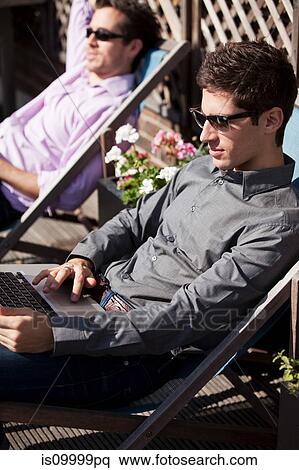 Stock Photography - Two men sunbathing on deck chairs with a laptop. Fotosearch
