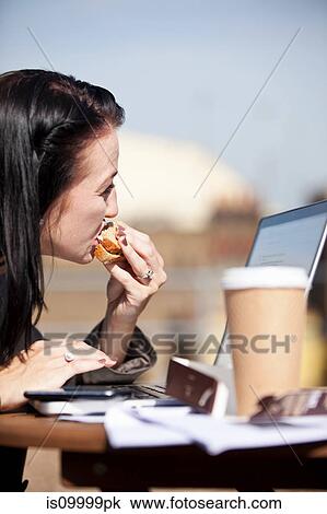 Stock Image - Young woman eating sandwich while using laptop. Fotosearch