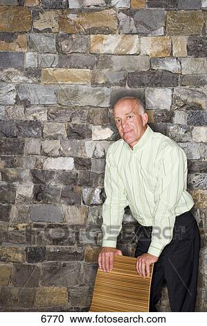 Mature Man Standing Beside Stone Wall Leaning On Chair Smiling