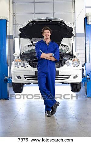 Male car mechanic, in blue overalls, standing in front of car with open